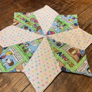May include: A star-shaped quilted trivet featuring panels of Peanuts comic strips and white fabric with pastel polka dots. The trivet has a light blue border and is designed to protect surfaces from heat.