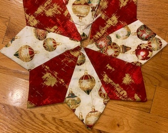 May include: A star-shaped Christmas tree skirt. The skirt is made of fabric with alternating red and white panels. The red panels have a gold brushstroke design. The white panels feature gold and red Christmas ornaments.
