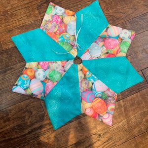 Op de afbeelding: Een stervormige kerstboomrok voor Pasen. De rok is gemaakt van stof met een patroon van kleurrijke paaseieren. De punten van de ster zijn effen turquoise met een glinsterende glans. Een turquoise lint is in het midden vastgebonden.