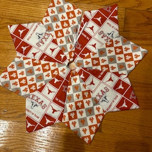 May include: A star-shaped table topper made of fabric with a checkered pattern in orange and gray. The fabric features the University of Texas logo and the words "Texas" and "The University of Texas".