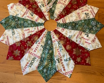 May include: A white, green, and red Christmas tree skirt with a snowflake and star pattern. The skirt has the words "Best Wishes", "Noel", "Happy Holidays", and "Season's Greetings" printed on it.