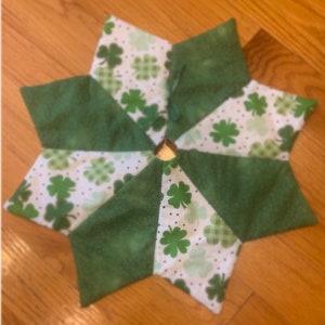 May include: A green and white patchwork tree skirt with a shamrock pattern. The skirt is made of fabric with a variety of textures and is perfect for a St. Patrick's Day celebration.