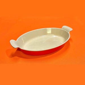 May include: Oval-shaped, red and white ceramic baking dish. The dish has a bright red exterior and a cream-colored interior. It features two white, ridged handles on either side. The dish is designed for baking and serving.