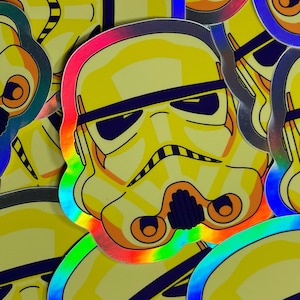 Peut inclure: Gros plan sur une pile d'autocollants holographiques représentant un casque de Stormtrooper de Star Wars en dessin animé jaune et noir. Les autocollants ont une bordure arc-en-ciel irisée.