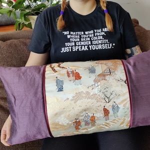 May include: A purple and cream pillow with a Japanese-inspired embroidered design featuring figures in traditional clothing and a landscape scene. The pillow has a burgundy trim. The text on the pillow reads: "NO MATTER WHO YOU ARE, WHERE YOU'RE FROM, YOUR SKIN COLOR, YOUR GENDER IDENTITY, JUST SPEAK YOURSELF."