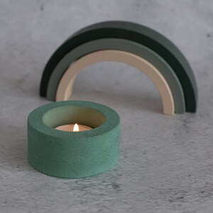 Puede incluir: Un portavelas de hormigón verde azulado con una pequeña vela blanca en su interior. El portavelas está sobre una superficie gris delante de un arcoíris de madera.