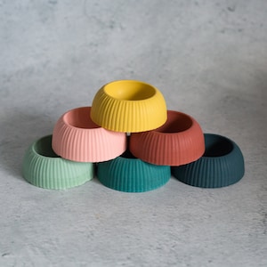 Puede incluir: Seis pequeñas macetas redondas y coloridas de hormigón. Las macetas están apiladas en forma de pirámide. Los colores son verde, amarillo, rosa, rojo y azul.