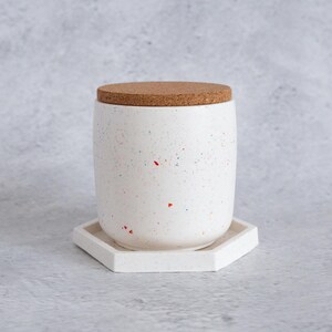 May include: White ceramic jar with a cork lid and a speckled pattern. The jar is sitting on a white hexagonal coaster.