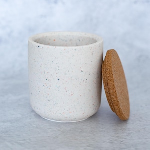 以下が含まれることがあります： コルクの蓋が付いた、斑点のある白いセラミック容器。容器は円筒形で、滑らかで丸みを帯びた縁があります。コルクの蓋は丸く、容器の側面に寄りかかっています。容器には小さな色の斑点があります。