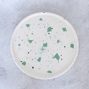 May include: A round, off-white ceramic plate with a speckled pattern. The plate features irregular, light green geometric shapes and small dark green speckles scattered across its surface. The plate is set against a light gray background.