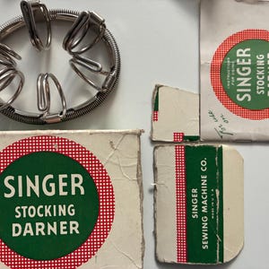 Puede incluir: Un surtido de accesorios de máquinas de coser Singer vintage. Incluye un zurcidor de medias de metal y varias cajas pequeñas y embalajes con el logotipo de Singer y el texto "Stocking Darner". El embalaje es blanco con un diseño circular rojo y verde.