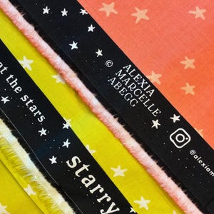 Può includere: Primo piano di tessuto colorato con un motivo a stelle. Il tessuto è nelle tonalità del giallo, rosa e arancione. Sono visibili nastri neri con testo bianco che recita "gaze at the stars" e "starry".