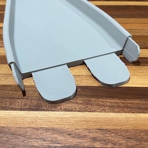 May include: A light gray 3D-printed ramp with two rounded extensions at the base. The ramp has a curved design with raised edges. The object is set against a wooden background.