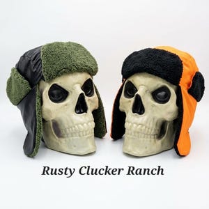May include: Two trapper hats displayed on skull models. The left hat is black with olive green faux fur lining. The right hat is bright orange with black faux fur lining. The text "Rusty Clucker Ranch" is printed below the two skull models.