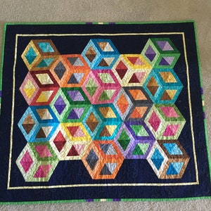 May include: A colorful quilt with a geometric design featuring interconnected, three-dimensional cube shapes in various colors. The quilt has a dark blue border with a green edge and a cream-colored inner border. The cube shapes include shades of blue, pink, orange, and green.