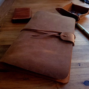 May include: A rectangular brown leather journal with a wrap-around leather tie closure. The journal features a smooth, matte finish and a single leather strap that secures the cover. The design is simple, focusing on the natural texture of the leather.