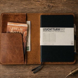 May include: A brown leather notebook cover holding a black Leuchtturm1917 notebook with a white paper band that reads LEUCHTTURM1917 DETAILS MAKE ALL THE DIFFERENCE. A Field Notes booklet is tucked into the left pocket, with a pen and inkwell nearby.