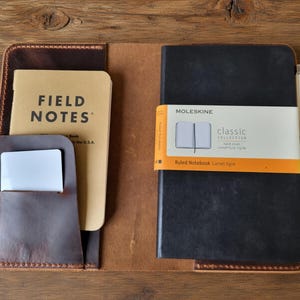 Op de afbeelding: Een bruine lederen notitieboekhoes met een linkerzak met een beige Field Notes notitieboekje en een witte kaart, en een rechterkant met een zwart Moleskine classic collection gelinieerd notitieboek met een witte en oranje band en een zilveren pen.
