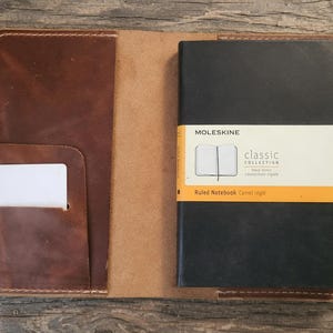 May include: A brown leather notebook cover featuring a left-side pocket with a white card, a black Moleskine classic collection ruled notebook on the right, and a yellow pencil held in a side loop, all stitched with light-colored thread.