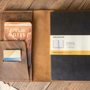 May include: A brown leather notebook cover containing a black Moleskine classic collection ruled notebook, a Field Notes booklet, a small card with a blue and gold design, and a silver pen held in a side loop, all displayed in an open position.