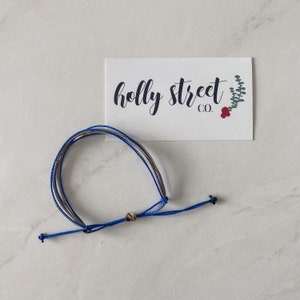 May include: A blue and tan bracelet with a small gold-colored bead. The bracelet is made of several strands of cord and is knotted at the ends. A white card with the text "holly street co." is in the background.