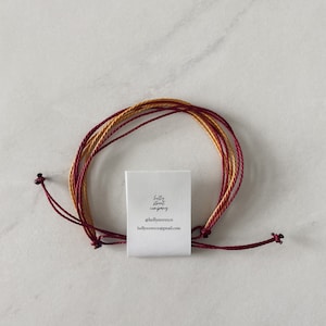 May include: A red and gold braided cord bracelet with a small white tag that reads "holly street company", "@hollystreetco", and "hollystreetco@gmail.com".