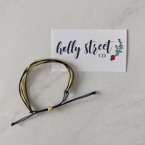 May include: A yellow and black friendship bracelet with a knot at the end. The bracelet is on a white surface with a business card that says "holly street co."