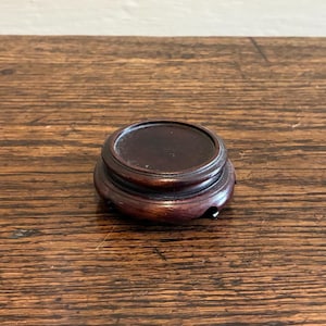 May include: A small, round, dark brown wooden stand with a raised, circular top. The stand has a tiered design and sits on a wooden surface. The stand is likely for displaying a small object.