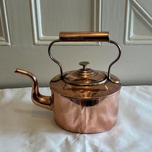 May include: A copper kettle with a curved spout and a handle. The kettle has a lid with a small knob and a curved handle. The kettle is sitting on a white surface. The kettle is made of copper and has a shiny finish.