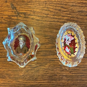May include: Two antique glass ashtrays with painted portraits. One is star-shaped with a portrait of a person in a military uniform. The other is oval-shaped with a portrait of a woman in a hat. Both have gold accents.