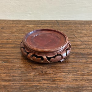 May include: A circular, dark brown wooden stand with a carved, decorative edge. The stand features a flat, circular top, designed to hold a small object. The wood grain is visible, enhancing its antique appearance.