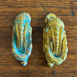 May include: Two antique brass door pulls with a gold finish and verdigris accents. Each pull features a decorative design with a central vertical element and a curved, leaf-like border. The pulls are set against a wooden surface.