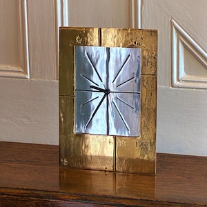 Könnte beinhalten: Eine rechteckige, goldfarbene Metalluhr mit silberfarbener Mitte. Das Zifferblatt hat ein Sternmuster. Die Uhr wird auf einer dunklen Holzoberfläche ausgestellt.