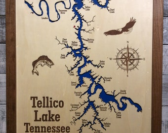 May include: A wooden map of Tellico Lake in Tennessee, with the lake outlined in blue and the surrounding land in brown. The map includes the names of various creeks and branches, as well as the town of Tellico Village. There is a compass rose in the upper right corner and a fish jumping out of the water in the lower left corner.