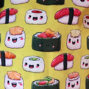 May include: A bright yellow fabric featuring a repeating pattern of various sushi pieces with cartoon faces. The sushi illustrations include nigiri with red fish, maki rolls with green avocado, and other rolls topped with orange fish, all with smiling expressions.
