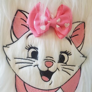 May include: Embroidered white cat face with pink accents and a pink bow with white hearts. The cat has black eyes, whiskers, and a pink nose. The bow is centered above the cat's head, creating a playful and whimsical design.