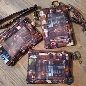 May include: Three brown and tan fabric wallets with a vintage library book design. One wallet has a wrist strap, another has a lanyard, and all have a clear ID window and a zipper closure. The wallets are on a wooden surface.