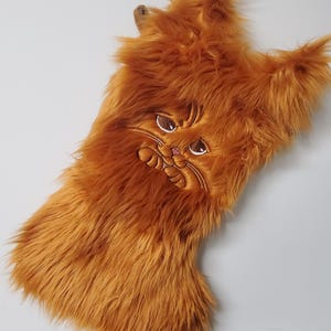 May include: A brown furry Christmas stocking with an embroidered cat face. The cat has pink cheeks and brown eyes.
