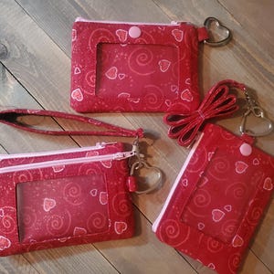 May include: Three red zippered pouches with clear plastic windows and pink hearts on a red and white swirl pattern. The pouches have a zipper closure and come with a wrist strap and a heart-shaped keychain.