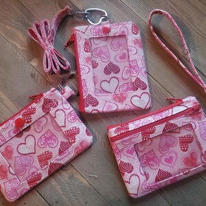 May include: Three pink fabric ID badge holders with a heart pattern. One has a lanyard, another a wrist strap, and the third a zipper. The holders have clear windows and red accents, set against a wooden background.