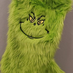 May include: Green fuzzy stocking with a Grinch face embroidered on the front. The Grinch's eyes are yellow and his mouth is a black stitched smile.