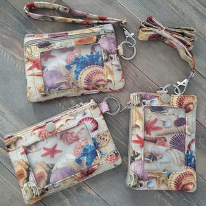 May include: Three fabric pouches featuring a seashell and starfish print in shades of tan, purple, red, and blue. Each pouch includes a clear plastic ID window, a metal zipper closure, and a detachable wrist strap or lanyard with a metal swivel clip.