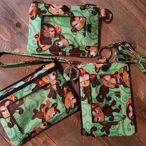 May include: Three fabric pouches with a light green background and a repeating pattern of brown monkeys with pink accents. Each pouch features a clear plastic window, a black zipper closure, and a metal key ring. One pouch includes a wrist strap.