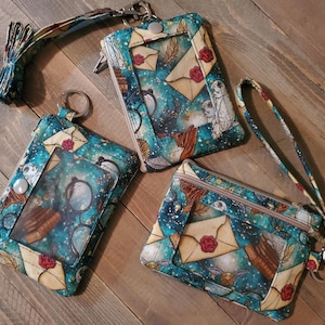 May include: Three fabric ID holders with a clear window, featuring a teal and gold celestial design with owls, letters, and glasses. One has a lanyard, another a wrist strap, and the third a keyring.