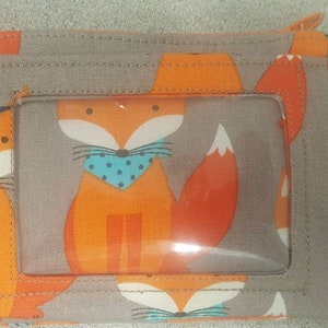 May include: A gray fabric zippered pouch with a clear plastic window. The pouch has a fox design with orange, blue, and white accents. The pouch has a metal ring attached to it.