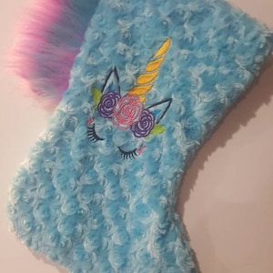May include: A light blue Christmas stocking with a unicorn design. The stocking has a soft, textured surface and is decorated with an embroidered unicorn face, a yellow horn, and a pink and purple mane.