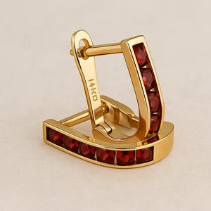 May include: A pair of gold-coloured hoop earrings. Each earring has a row of small, red gemstones set along the outer edge. The earrings are marked with "14K Gold".