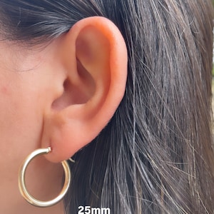Puede incluir: Pendientes de aro de oro de 25 mm de di&aacute;metro. Los pendientes se muestran en la oreja de una persona.
