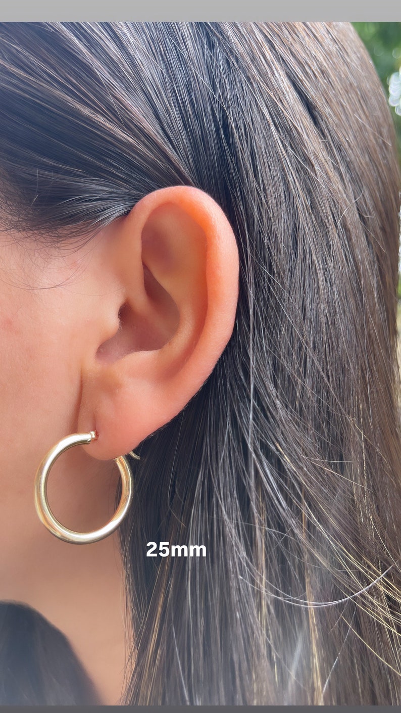 Puede incluir: Pendientes de aro de oro de 25 mm de di&aacute;metro. Los pendientes se muestran en la oreja de una persona.