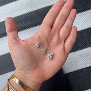 May include: Two gold pendants with clear round stones, one larger than the other, are displayed on a hand.